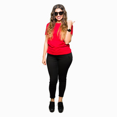 Young beautiful woman wearing red t-shirt and sunglasses smiling with happy face looking and pointing to the side with thumb up.