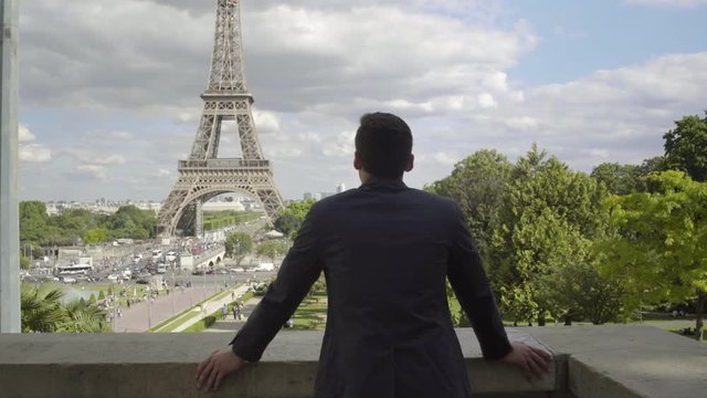 Businessman posing at the Eiffel Tower after work, with a white shirt, a jacket over his shoulder, brown hair and tanned skin. Combing his hair. Beautiful cloudy blue sky in Paris. 4K. Tracking in.