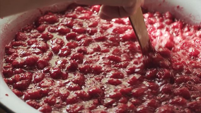 Female hand stirring raspberry jam in an enameled bowl with a wooden spatula.