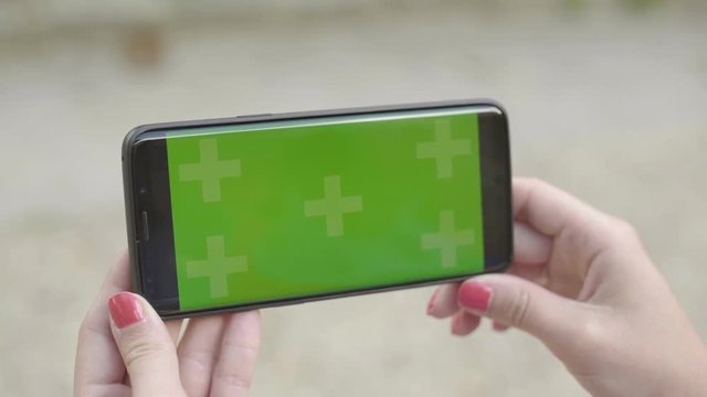 Close-up of smartphone with Green Screen Sitting on bench in park, holding by woman in Horizontal Landscape Mode, watching movie on touch screen with Tracking Markers. Watching Content, Videos, Blogs.