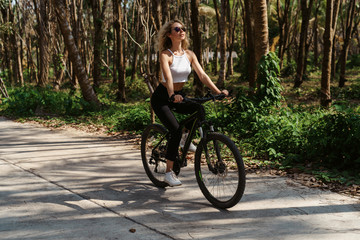 Fototapeta premium Pretty woman with Bicycle on a background of palm trees forest. Young girl riding bike in Vietnam or Thailand tropical jungle with coconut palms. Summer holiday tourist activity. Active sports tourism