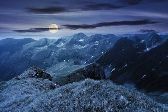 beautiful landscape of fagaras mountains at night in full moon light. rocks on steep grassy slopes. snow in the deep valley. wonderful summer weather with clouds on the sky