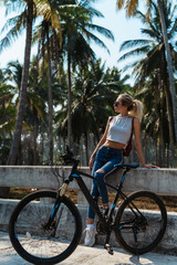 Naklejka premium Pretty woman with Bicycle on a background of palm trees forest. Young girl riding bike in Vietnam or Thailand tropical jungle with coconut palms. Summer holiday tourist activity. Active sports tourism