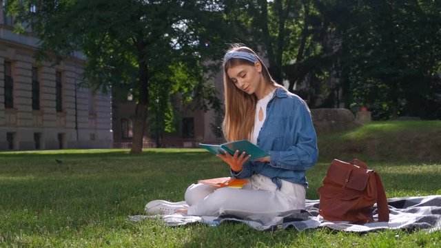 Cheerful student girl studying her notes. Young woman sitting on the grass in the park, holding an open notebook, looking away and smiling. Happy student concept.