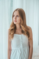 Girl in a light white dress posing by the window.