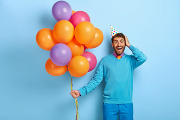 Studio shot of excited joyful man looks with great happiness at camera, wears blue clothes comes on festive event glad to meet with friends holds bunch of multicolored helium balloons. Holiday concept