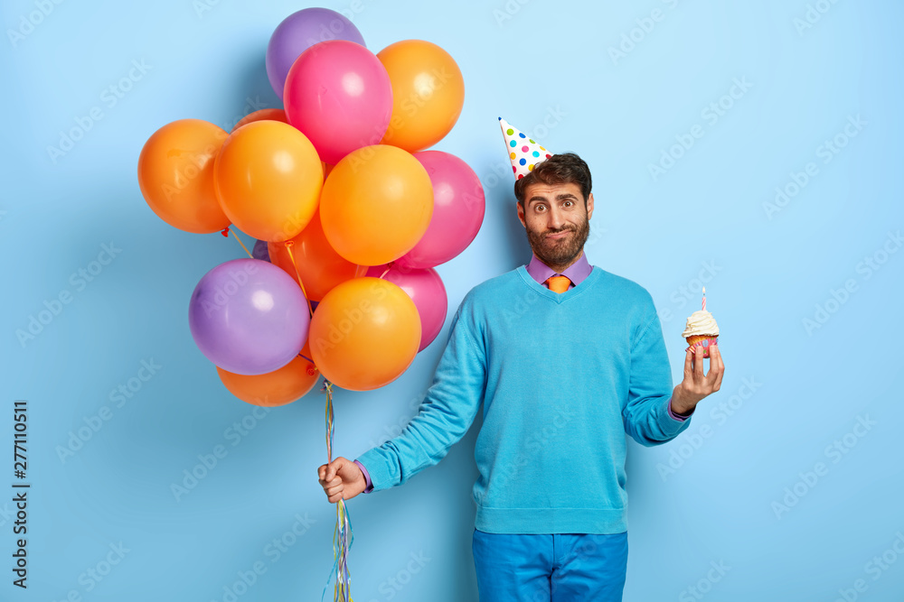 Wall mural horizontal shot of friendly guy with puzzled look, stands with cupckae and balloons, celebrates anni