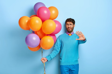 Dissatisfied man celebrates anniversary of birth, shows refusal gesture, doesnt want to be photographed, frowns face, wears spectacles, poses with colorful balloons. Guy with party attributes