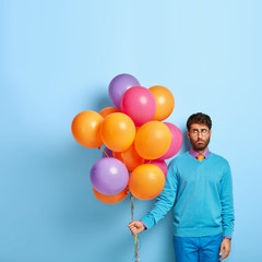 Fototapeta na wymiar Displeased balloon artist stands with party accessories, has sad expression, tired after decoration for holiday, wears blue outfit, stands indoor. Gloomy elegant birthday man being in low spirit