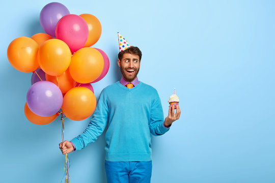 Young father in festive outfit, celebrates first birthday of his son, holds muffin with burning candle, colorful balloons, has joyful surprised look, isolated on blue wall, free space for text