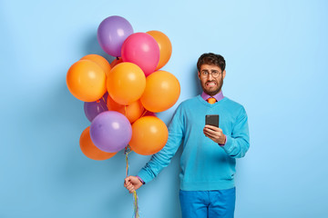 Dissatisfied young man organises holiday on special occasion, unhappy to read bad news in internet, holds mobile phone, has spoiled party, finds out about tragedy with friend carries colorful balloons