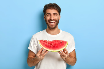 Waist up shot of smiling man enjoys summer day, holds slice of fresh ripe watermelon, wants to eat...