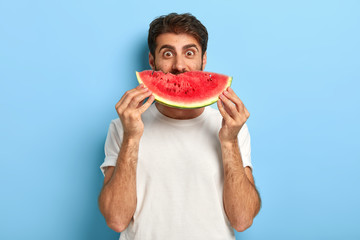 Funny man holds half of red watermelon near face, has glad surprised look, dressed casually, has healthy summer nutrition. European guy poses with tasty tropical fruit against blue background