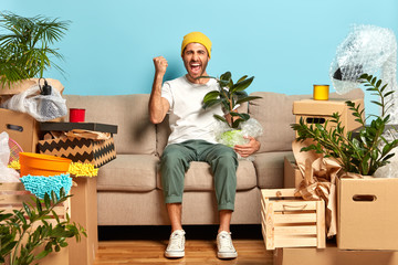 Shot of overjoyed guy clenches fist, poses at sofa, holds wrapped houseplant, moves in new modern...