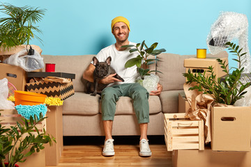 Indoor shot of delighted carefree house owner sits on couch and hugs pet, rests in own apartment,...