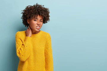 Beautiful unhappy dark skinned woman suffers from neck ache injury, has dissatisfied facial expression, wears yellow jumper, poses indoor against blue wall with blank space for your information