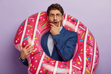 Embarrassed shocked boss holds breath from fear, stares at camera, poses with rubber swimring,...