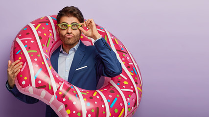 Fashionable male entrepreneur wears goggles, elegant suit, carries inflated rubber ring, needs vacation and rest from office work, prepares for summer holidays, isolated on violet background