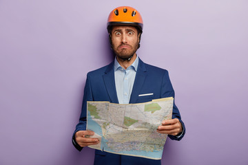 Photo of embarrassed male architect learns location for building, holds paper map, wears headgear,...