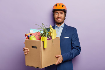 Business and new job concept. Displeased office worker carries heavy cardboard box with personal...