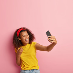 Naklejka premium Smiling African American woman has video conversation with friend, looks hapily at smartphone camera, points with index finger, dressed casually, models over pink wall blank copy space above