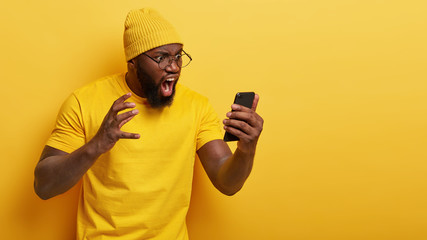 Outraged annoyed dark skinned man stares angrily at smart phone screen, shouts with irritation,...