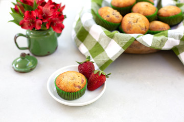 Obraz na płótnie Canvas Freshly Baked Strawberry Muffins in wooden bowl with one isolated in front on plate; green and white checked cloth lining bowl; red flowers in green tin.
