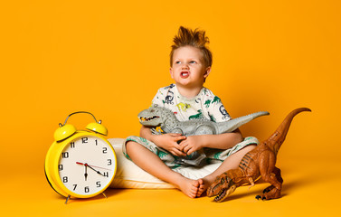 Little cute boy in pajamas holding a toy dinosaur in his hands, sitting on a pillow with an alarm...