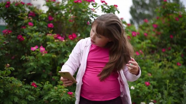European girl with blonde hair taking pictures of herself on her smartphone. Girl takes a selfie on the phone in the garden on a background of blooming roses .