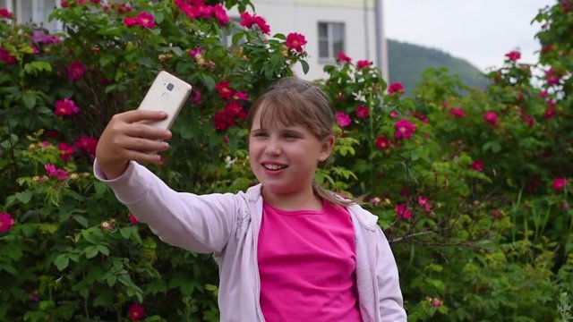 European girl with blonde hair taking pictures of herself on her smartphone. Girl takes a selfie on the phone in the garden on a background of blooming roses .
