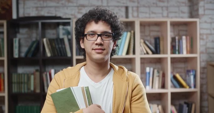 Asian guy with curly hair is standing between bookshelves of library holding books in hands, looking at camera and smoling - portrait closeup 4k, education, student lifestyle concept 4k