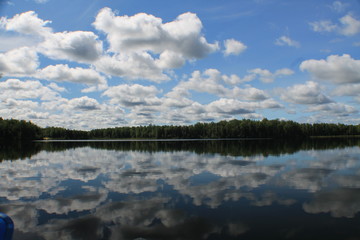 Blue sky w scattered cumulus clouds reflecting on a still tree-lined lake