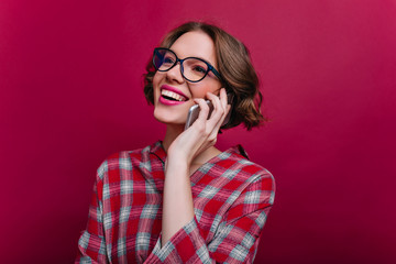 Close-up portrait of blissful brunette girl with curly hairstyle talking on phone. Attractive caucasian lady in glasses and checkered shirt posing with smartphone on claret background.