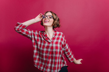 Fototapeta premium Pretty brunette girl in casual shirt expressing positive emotions on claret background. Studio shot of cheerful curly young lady in glasses.