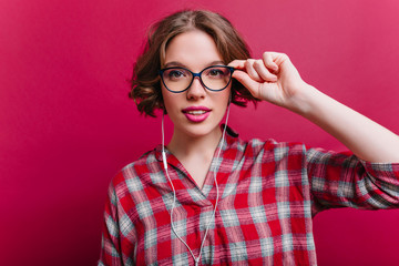 Sensual young woman with pink lips looking with interest and touching glasses. Studio shot of charming curly girl with tattoo posing in white earphones.