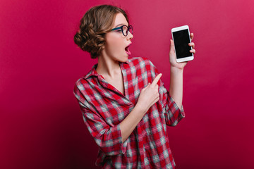 Emotional white girl with curly hairstyle posing with smartphone on claret background. Indoor portrait of amazed stylish woman in casual attire holding cell.