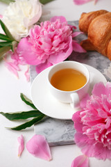 cup of tea herbal, croissant and peony buds on marble plate on white table. Beauty breakfast composition, celebration seasonal holiday feminine background