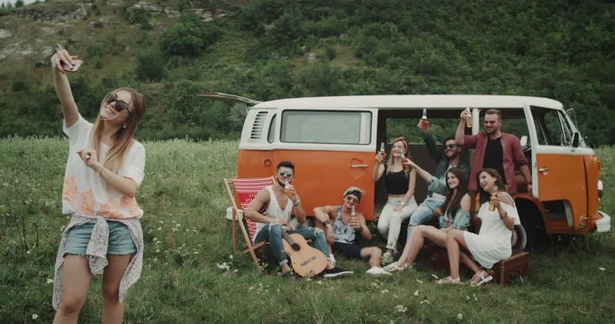 Selfie time for a big group of friends at the picnic party taking pictures from phone , they are sitting beside of the retro van , in the middle of landscape