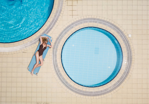 Beautiful and sexy woman have sunbathe in brown swimmsuit next to round pools, laying on a blue sunbed. Aerial model image, from birds eye view, by drone.