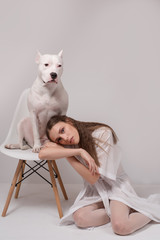 Fashionable young woman in white dress sitting on the floor and touching her lovely white staffordshire dog sitting on chair, in front of white background. Close relationship between human and dog