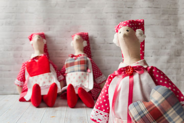 Obraz premium Three tilde dolls in red dresses, and white aprons, and red kalpaks sit on a light background. Interior dolls. There is a place for text. The concept of needlework,.