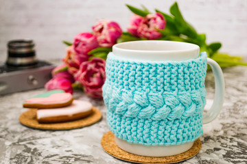 A white cup of tea in a knitted blue case with an old camera and pink flowers tulips and sweets, gingerbread on a textural table.