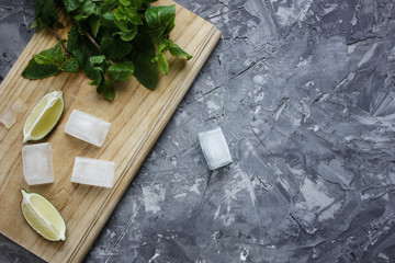 Ingredients for cocktail mojito. Fresh lime pieces, mint and ice on the wooden cutting board on the grey concrete background with cracks and scratches