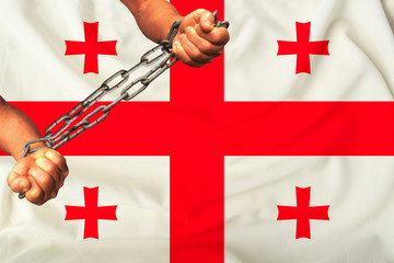 men's hands chained in heavy iron chains against the background of the flag of Georgia on a gentle...