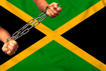 men's hands chained in heavy iron chains against the background of the flag of Jamaica on a gentle...