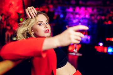 Photo of young blonde in red suit looking at camera sitting at bar with cocktail glass in nightclub