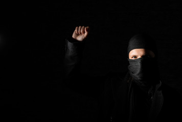 Young adult girl in black clothes with hidden face protests raising fist on black background with...