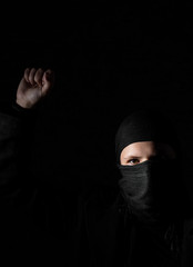 Young adult girl in black clothes with hidden face protests raising fist on black background with...