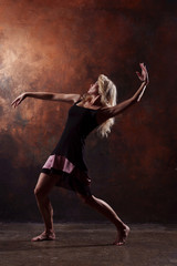 Fototapeta na wymiar Photo of young dancing blonde looking back in black dress on brown background