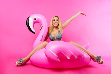 Photo of happy blonde in bathing suit and pink glasses with raised hand up sitting on inflatable flamingo on empty pink background